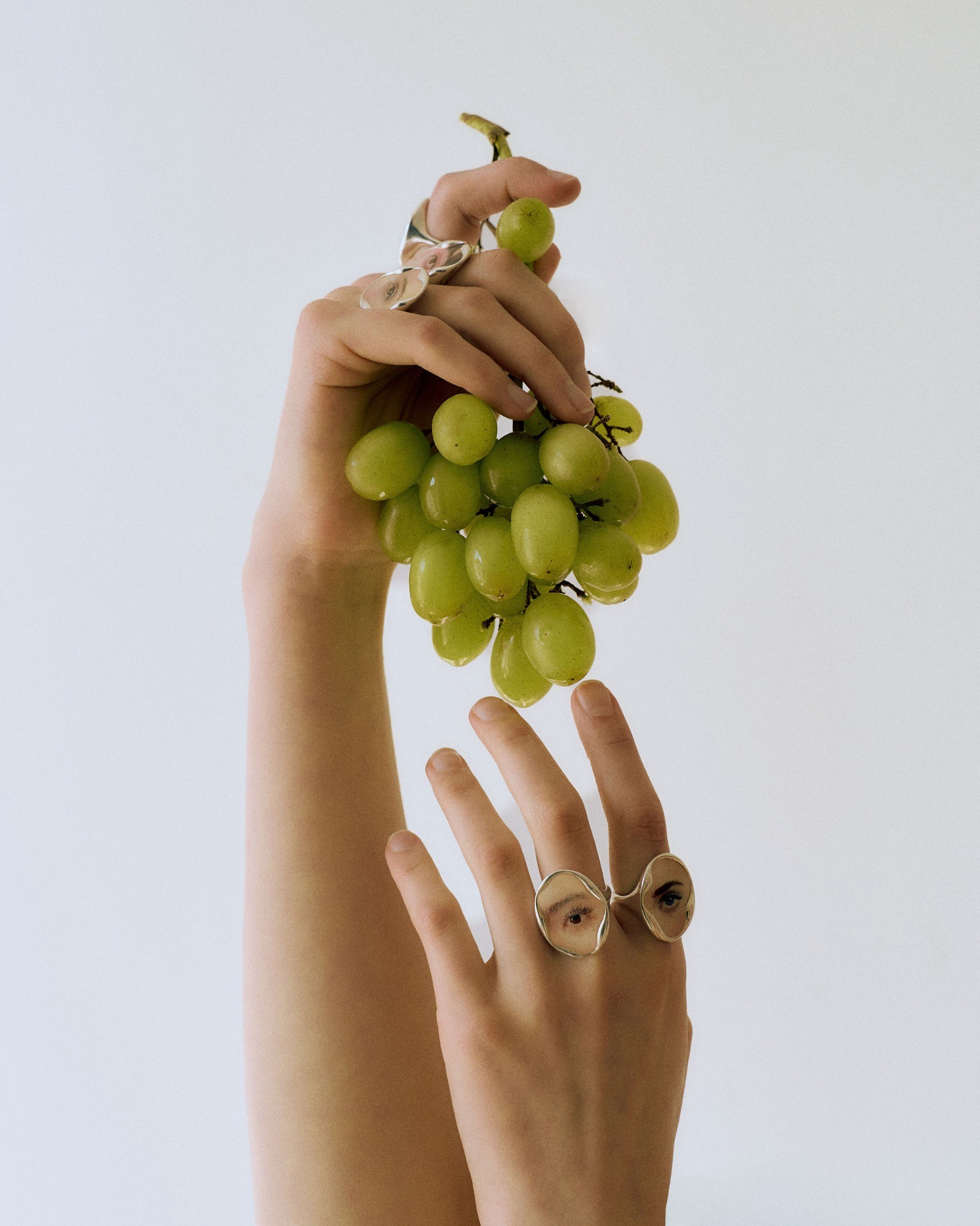 Hands wearing silver eye portrait rings and holding green grapes